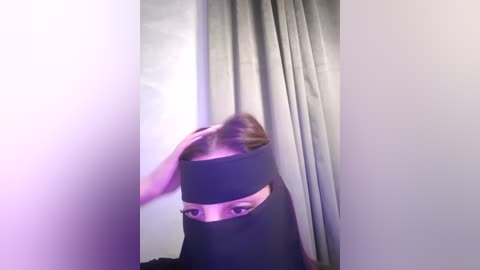 Media: Video of a woman in a black headscarf with her hair pulled back, standing in a dimly lit room with gray curtains. The image is slightly blurred, giving a soft, dreamy effect.