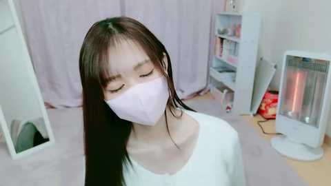 Media: Video of an East Asian woman with long, straight black hair, wearing a white mask, white top, and pinkish-purple curtains in a cozy, minimalistic room with a white bookshelf and a heater.