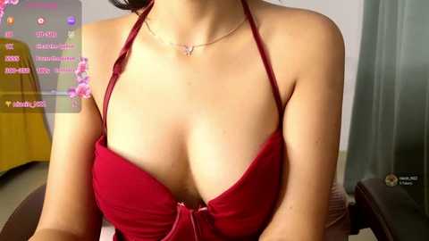 Video of a light-skinned woman with a slender physique, wearing a red halterneck dress that reveals ample cleavage, and a dainty necklace. Background features a yellow couch and green glass door.