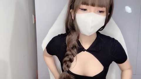 Media: Video of an Asian woman with long brown hair in a braid, wearing a black crop top revealing cleavage, white face mask, and white chair background.