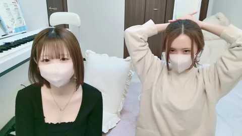 Media: A video shows two Asian women with light skin, brown hair, and surgical masks, one sitting at a piano, the other stretching, in a modern, white-tiled room.