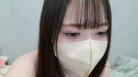 Media: Video of a young Asian woman with straight black hair and bangs, wearing a beige surgical mask, looking down. Background shows a blurred, light-colored wall.