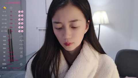 Media: Video of a young Asian woman with long black hair, wearing a white fluffy bathrobe, looking down, in a modern, softly lit bedroom with a white lamp.