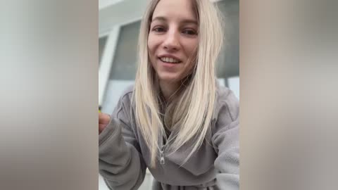 Media: Video of a smiling young woman with long, straight blonde hair, wearing a gray hoodie, standing in a modern, well-lit room with glass doors.