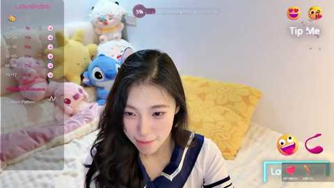 Media: Video of a young Asian woman with long black hair, wearing a white sailor uniform, sitting on a bed with stuffed animals. Background shows a cluttered room with a blue and yellow patterned blanket.