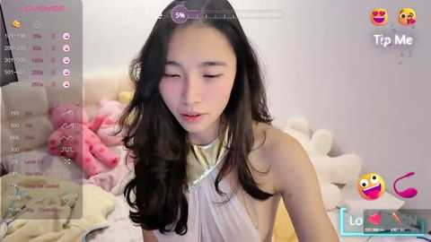 Media: A young Asian woman with long black hair and fair skin, wearing a sleeveless white dress, sits on a bed with pink pillows, appearing to be on a video call.