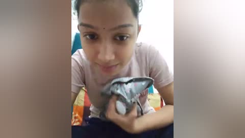 Media: Video of a young South Asian girl with medium brown skin, short black hair, wearing a light pink t-shirt and holding a white and blue toy car. She is sitting on a colorful blanket in a well-lit room.