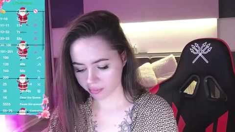 Media: Video of a young woman with long brown hair, wearing a black and white patterned top, sitting in a red and black gaming chair. Background shows a green screen with Twitch streamer statistics and a stuffed animal.