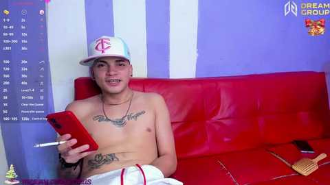 Media: Video of a shirtless young man with tattoos, wearing a white baseball cap, holding a red phone, lounging on a red couch in a purple-striped room.