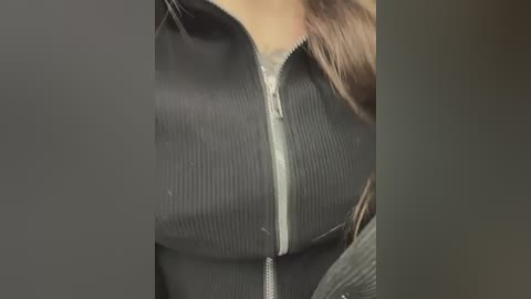 Media: Video of a woman's torso wearing a black, ribbed zip-up jacket with a visible white zipper, partially unzipped to reveal a grey sweater underneath. Her long, brown hair is partially visible.