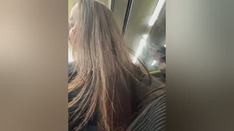 Media: A video captures a young woman with long, straight brown hair, partially covering her face, sitting in a dimly lit bus. She wears a dark, textured sweater. The background shows reflections and indistinct shapes.