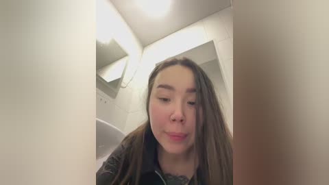 Media: Video of a young woman with light skin and long brown hair, wearing a green jacket, looking down into a white toilet stall.