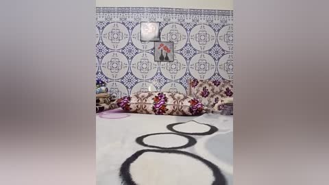 Media: Video of a bedroom with white walls and blue and white tiled backsplash. A bed covered in a floral duvet and pillows, adorned with a black abstract rug.