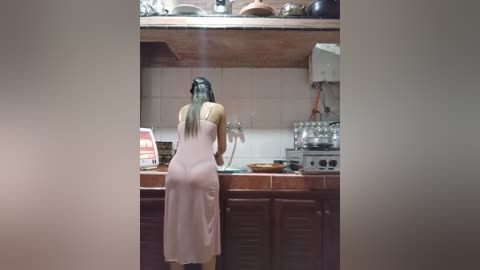 Media: Video of a woman in a pink apron and long black hair, cooking in a rustic kitchen with wooden cabinets and a tiled backsplash.