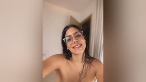 Media: A video of a smiling woman with long, dark hair, wearing glasses, and a white top, standing in a dimly lit room with cream-colored walls.