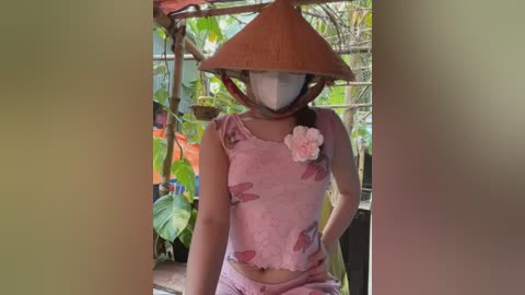 Media: Video of a person wearing a pink, floral-patterned outfit and a large, conical hat, with a white mask covering the face, standing in a lush, green garden filled with hanging plants and colorful flowers.