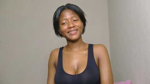 Media: Video of a smiling African woman with short black hair, wearing a black tank top, against a plain beige wall.