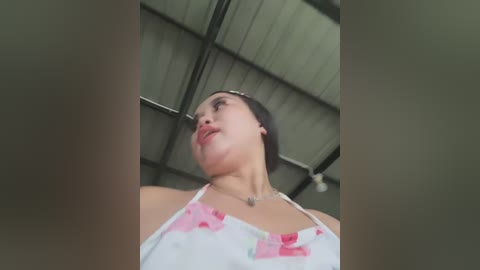 Media: A low-angle video of a woman with fair skin and dark hair, wearing a white dress with pink floral patterns, standing in a warehouse with a metal ceiling.