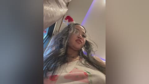 Media: Video of a young woman with long, wavy, silver hair and fair skin, wearing a white shirt with red cherries, in a dimly lit room with a festive red hat and Christmas decorations.