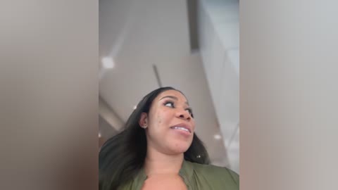 Media: A video of a light-skinned Black woman with long, straight black hair and a medium complexion, wearing a green top, looking upward with a slight smile. The background features blurred, neutral-toned walls and a staircase.