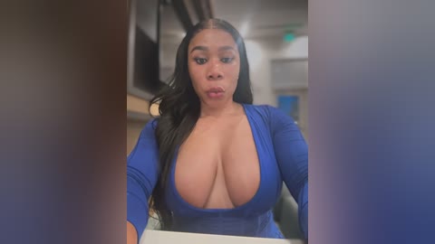 Media: Video of a curvaceous, dark-skinned woman with long, wavy black hair, wearing a low-cut, blue, long-sleeve dress, revealing ample cleavage, standing in a modern, neutral-toned room with blurred background.