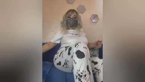 Video of a woman with light skin and blonde hair, wearing a cow-patterned bodysuit and mask, seated on a blue couch, against a beige wall with hexagonal decorations.