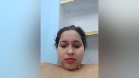 Media: Video of a South Asian woman with medium brown skin, black hair tied back, wearing red lipstick, and a gold necklace. She is indoors against a light blue wall with a white shelf and yellow trim.