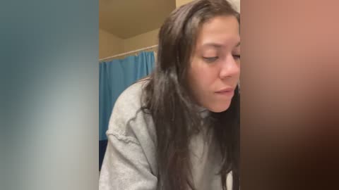 Media: Video of a young woman with long, dark hair, wearing a gray hoodie, leaning against a brown wall in a bathroom with a blue shower curtain.