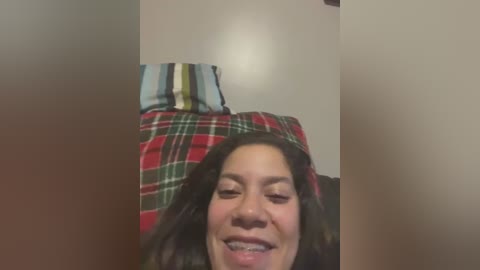 Media: Video of a smiling, light-skinned woman with dark hair, wearing a red plaid shirt, with a blurry background featuring a striped pillow.