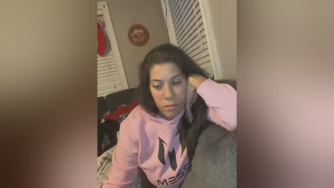 Media: Video of a young woman with long dark hair, wearing a pink hoodie, resting her head on a grey couch, in a dimly lit room with white shutters and red and white decorations.