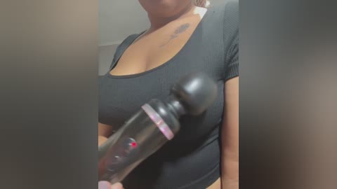 Media: Video of a woman with medium skin tone, wearing a black ribbed short-sleeve top, holding a black vibrator with a pink base, blurred background.