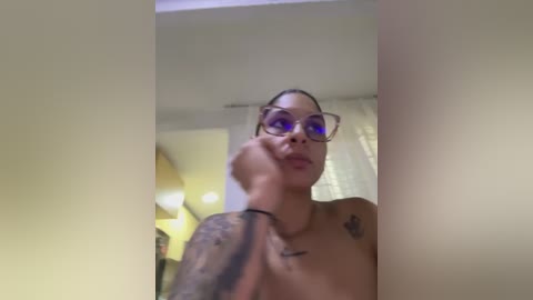 Media: Video of a topless, light-skinned woman with tattoos on her chest, wearing glasses, in a dimly lit room with a white curtain.