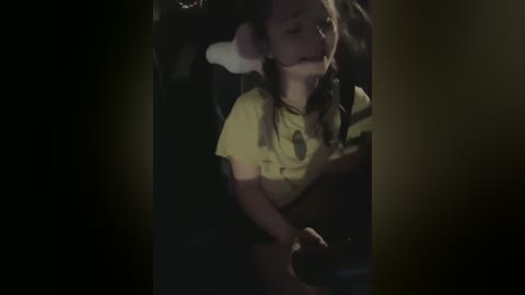 Media: Video of a young girl, possibly Caucasian, with long dark hair, wearing a yellow t-shirt, seated in a dark car. She appears relaxed, eyes closed, with a white pillow on her head.