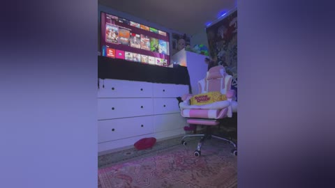 Media: Video of a dimly-lit, cozy gaming room with a large TV displaying a game interface, a white dresser, and a pink gaming chair with a \"KFC\" blanket, under a purple light.