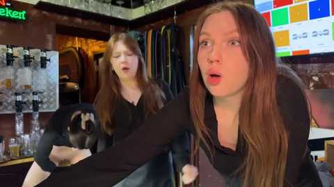 Media: Video of a young redhead woman in a black top, shocked expression, looking at a dog in a mirror, in a cluttered room with colorful posters, a keg, and clothes.