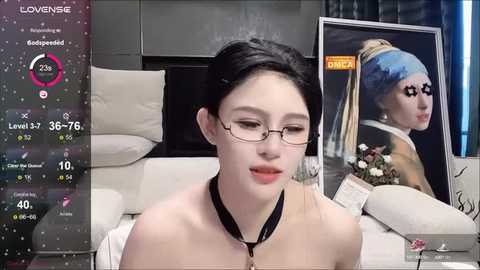 Media: Video of an Asian woman with pale skin, black hair, and glasses, wearing a beige top, sitting in a modern room with a painting of \"Girl with a Pearl Earring\" on the wall.