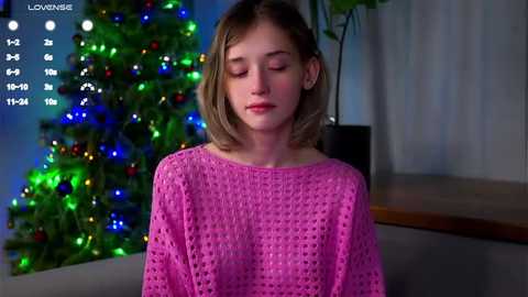 Media: A video of a young girl with blonde hair and fair skin, wearing a pink crocheted sweater, standing in front of a lit Christmas tree with colorful lights, indoors.