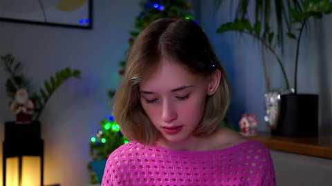 Video of a young woman with light skin and straight, shoulder-length blonde hair, wearing a pink knitted sweater. Background includes a Christmas tree, potted plants, and festive lights, creating a cozy indoor setting.