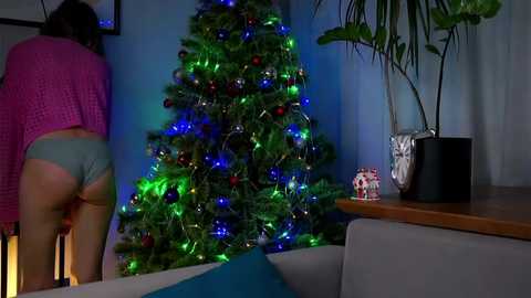 Media: Video of a woman in a pink sweater bending over to decorate a Christmas tree with multicolored lights. Background features a modern living room with plants and a wooden table.