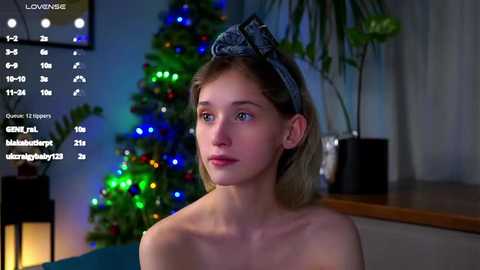 Media: Video of a young woman with fair skin, blue eyes, and blonde hair wearing a blue headband, looking thoughtful in a dimly lit room with a Christmas tree, colorful lights, and a potted plant in the background.