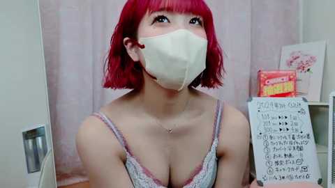 Media: Video of a woman with red bob hair, wearing a white face mask, pink bra, and a signboard with Japanese text in a room with pink curtains and floral decor.