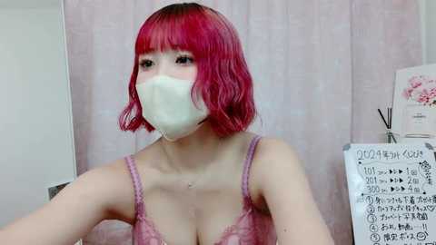 Media: Video of a young woman with bright red hair and a white face mask, wearing a pink lace bra, against a pink floral background.