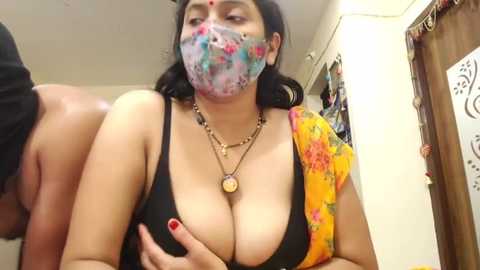 Media: Video of a South Asian woman with medium skin tone, wearing a floral yellow sari, black bra, and face mask, partially exposed breasts, in a cluttered, dimly-lit room.