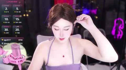 Media: Video of a young Asian woman with light skin and brown hair, wearing a purple spaghetti-strap top, adjusting her hair. Background features a dimly lit room with pink neon lights and a pink microphone stand.