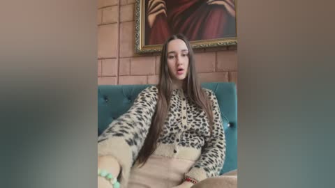 Media: Video of a young woman with long brown hair, wearing a leopard-print cardigan and beige skirt, sitting on a teal tufted sofa in a room with a pink brick wall and a framed painting of a person.