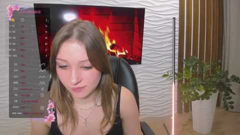 Media: Video of a young Caucasian woman with light skin, shoulder-length brown hair, wearing a black tank top, sitting in front of a lit fireplace. Calendar, potted plant, and floral decorations in background.