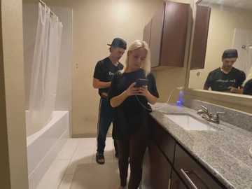 Media: Video of a modern bathroom with a blonde woman in a black top and jeans, standing by a granite countertop, while a man in a black cap and shirt looks at her.