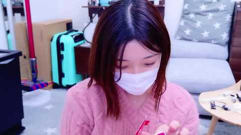 Media: Video of an East Asian woman with shoulder-length dark hair, wearing a pink sweater and white face mask, sitting indoors, holding a red pen. Background includes a gray chair, boxes, and a colorful toy.