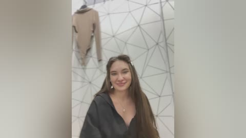 Media: Video of a smiling young woman with long brown hair, wearing a black hoodie, standing in a store with geometric patterned wall and hanging beige jacket.