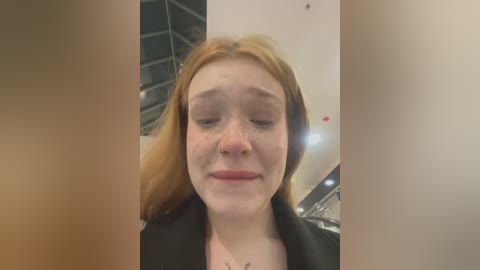 Media: Video of a young Caucasian woman with fair skin and reddish-brown hair, wearing a black top. She has a slight smile, with closed eyes and a relaxed expression. Background shows a modern interior with glass panels and white walls.